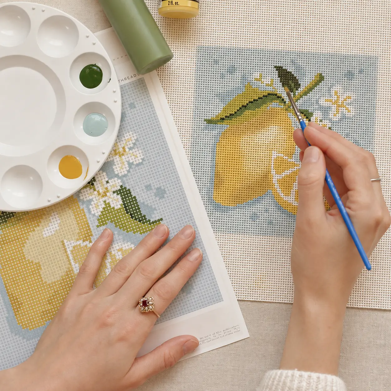 Hands painting a lemon needlepoint design onto mono canvas from a gridded color chart.